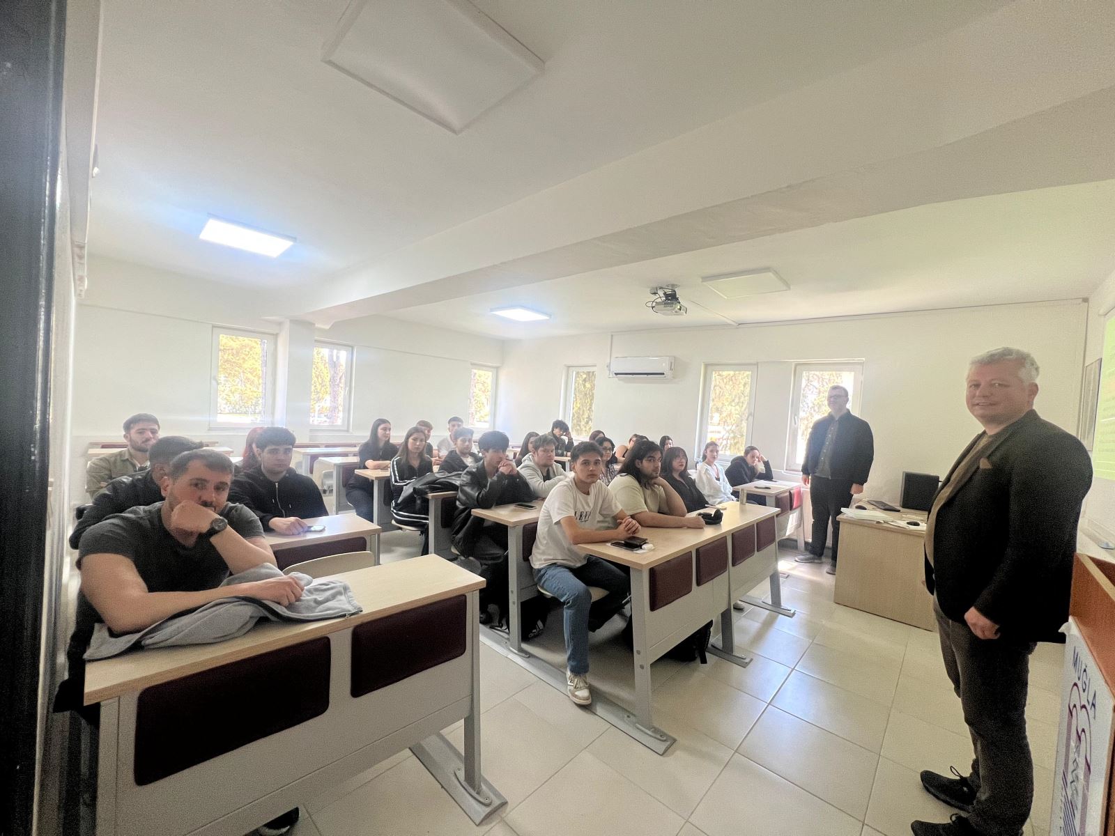 During a classroom meeting, students are seated at their desks and facing the camera. One speaker stands in the foreground while another stands near the students at the front of the room; a projection screen and large windows are visible in the classroom.